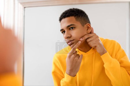 Foto de Adolescente chico en sudadera con capucha amarilla inspecciona cuidadosamente su piel facial para el acné, mostrando expresión enfocada en el espejo en habitación luminosa, teniendo problemas con la piel - Imagen libre de derechos