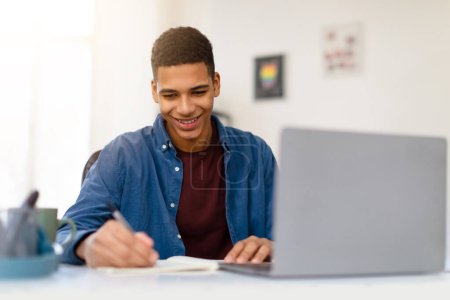 Foto de Sonriente chico adolescente negro en camisa azul toma notas de la computadora portátil, disfrutando de una sesión de estudio productiva en casa en un espacio de trabajo brillante y organizado - Imagen libre de derechos