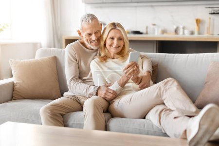 Senior couple sitting closely on their couch, sharing a joyful moment while looking at a smartphone in a cozy home