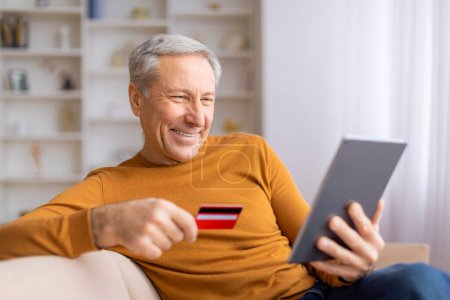Foto de Un hombre mayor sonriente sostiene una tarjeta de crédito mientras usa una tableta para comprar en línea desde su sofá, mostrando conveniencia - Imagen libre de derechos
