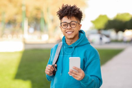 Photo for A joyous young brazilian guy in blue hoodie smiling while using a phone with a park backdrop - Royalty Free Image
