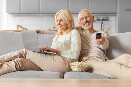Téléchargez les photos : Couple sénior multi-tâches avec ordinateur portable et télécommande, passer une soirée détendue à la maison - en image libre de droit