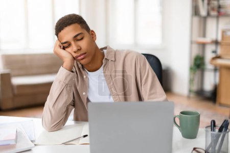 Téléchargez les photos : Un jeune homme semble fatigué et ennuyé avec sa tête reposant sur sa main alors qu'il est assis à un bureau avec un ordinateur portable et du matériel d'étude - en image libre de droit