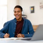A joyful young man smiles while listening to music through white headphones and taking notes with a pen