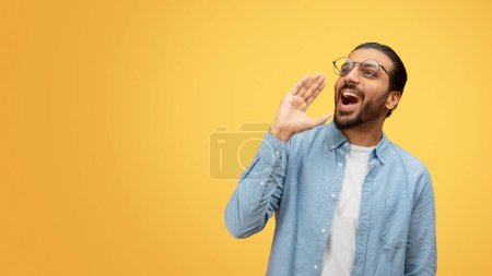 A vocal indian man uses his hand as a megaphone to shout something out against a yellow background