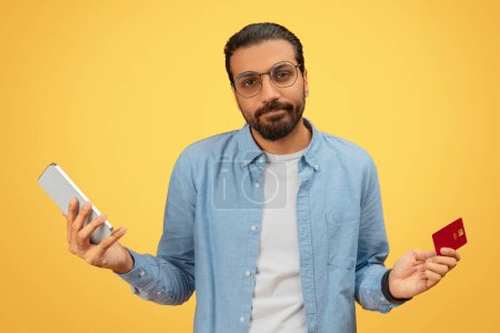 Téléchargez les photos : Un Indien déçu en chemise bleue affiche un téléphone portable d'une main et une carte de crédit rouge de l'autre, sur fond jaune - en image libre de droit