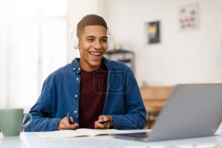 Smiling teenage guy in headphones playfully imitates drum playing in front of his laptop, showing creativity and enjoyment while engaging with digital content