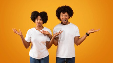 Shocked african american couple gesturing and grimacing on yellow studio background. Appalled black man and woman looking at camera with confusion face expression, panorama
