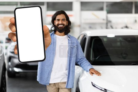 Foto de Hombre indio sonriente sosteniendo un teléfono con pantalla en blanco frente a él en un concesionario de automóviles, listo para la marca - Imagen libre de derechos