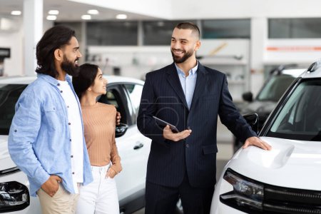 A car dealership interaction where a salesman smiles while discussing with indian couple beside a vehicle