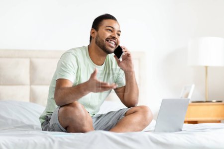 Téléchargez les photos : Un homme afro-américain joyeux s'assoit sur un lit avec un ordinateur portable, engageant une conversation sur son téléphone portable, dépeignant un cadre de travail détendu de la maison - en image libre de droit