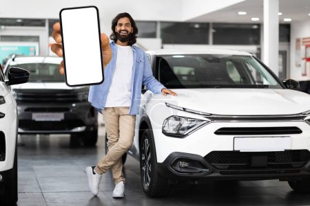 Téléchargez les photos : Un homme hindou joyeux aux cheveux longs tend un téléphone avec un écran vide dans une salle d'exposition de voiture, avec de nouvelles voitures en arrière-plan - en image libre de droit
