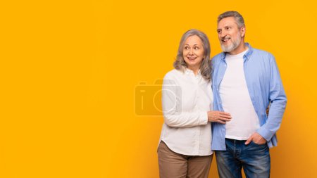 Photo for Happy Caucasian senior couple hugging while looking aside at empty space for sale offer, posing on yellow banner together, studio shot of mature spouses advertising service or product. Panorama - Royalty Free Image