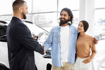 Junges glückliches indisches Paar beim Händeschütteln mit einem Verkäufer in einem Autohaus, der über einen Neuwagenkauf nachdenkt
