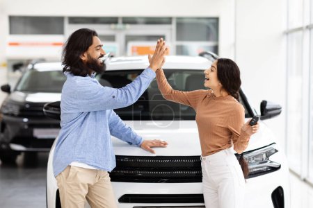 Téléchargez les photos : Un couple indien joyeux célèbre l'achat d'une nouvelle voiture avec un high-five dans un showroom concessionnaire, mettant en valeur leur bonheur et leur partenariat dans ce moment significatif - en image libre de droit