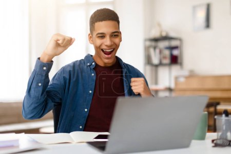 Foto de Exuberante chico adolescente negro aplaude con el puño bomba mientras mira a su computadora portátil, signo de triunfo y emoción en el logro de la meta personal o la victoria - Imagen libre de derechos