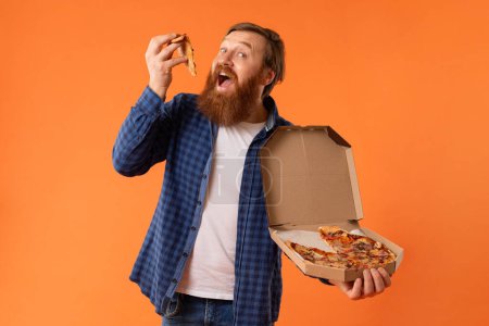 Téléchargez les photos : Homme barbu aux cheveux roux affamé tenant la boîte de livraison et dégustant une tranche de pizza, satisfait ses envies de malbouffe, sur fond de studio orange. Concept de mauvaises habitudes alimentaires, nourriture réconfortante - en image libre de droit