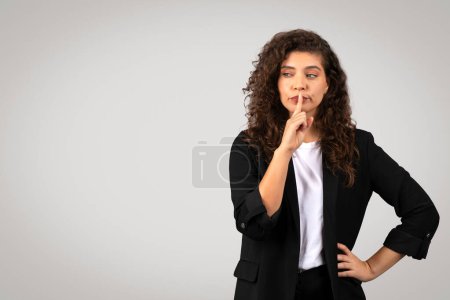 A curious woman in a smart blazer gesturing silence with her finger on the lips, depicting secrecy or quiet concept on a plain background