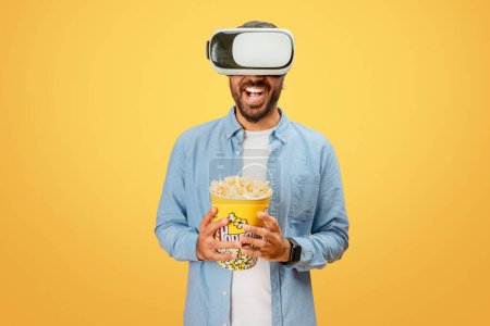 Ein von Virtual Reality faszinierter indischer Mann hält Popcorn in der Hand und genießt ein immersives digitales Erlebnis auf gelbem Hintergrund
