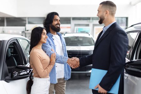 Joven pareja india estrechando la mano con un vendedor de coches en una sala de exposición de concesionarios, que simboliza una compra de coche nuevo