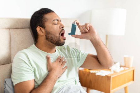Un homme afro-américain assis sur le lit utilise un inhalateur pour soulager une crise d'asthme, avec des tissus et de l'eau sur une table d'appoint