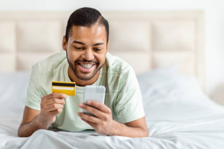 Téléchargez les photos : Homme noir souriant utilisant carte de crédit et smartphone, concept de shopping en ligne ou de banque à domicile - en image libre de droit