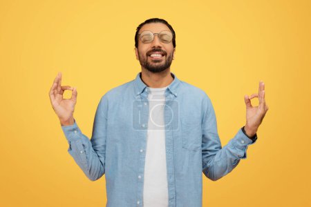 Joyful bearded indian man in glasses makes a peace sign with both hands on a yellow background