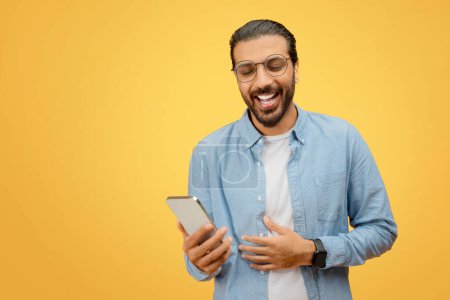 Foto de Un hombre indio alegre en gafas se ríe mientras mira su teléfono inteligente, vestido con una camisa de mezclilla casual - Imagen libre de derechos
