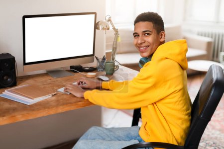 A happy young man in a yellow hoodie sits at his desk with a blank computer screen, ready to work or study