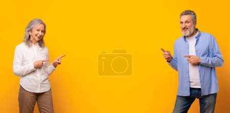 Photo for Happy mature spouses pointing fingers at middle of the banner, showing copy space between them, standing over yellow studio wall, smiling to camera while recommending nice deal. Panorama - Royalty Free Image