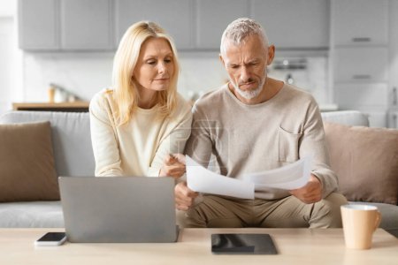 Elderly couple analyzing papers with attention, using a laptop and tablet for reference at home