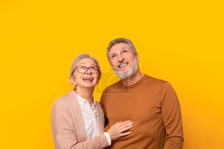 Téléchargez les photos : Couple âgé joyeux posant ensemble étreignant et regardant vers le haut sur fond de studio jaune, rêvant d'une vie à la retraite heureuse, annonçant une offre agréable pour les personnes âgées. Espace de copie - en image libre de droit