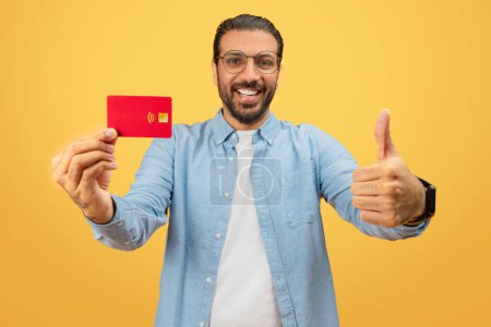Téléchargez les photos : Un homme indien joyeux en tenue décontractée montre une carte de crédit rouge à la caméra, donnant un pouce vers le haut avec un geste d'approbation - en image libre de droit