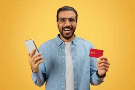 Foto de Un indio barbudo extático sostiene un teléfono inteligente y una tarjeta de crédito, lo que sugiere una transacción exitosa con un fondo amarillo - Imagen libre de derechos