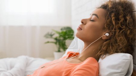 Téléchargez les photos : Femme afro-américaine écoutant de la musique calme et relaxante, couchée au lit - en image libre de droit