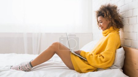 Foto de Mujer afroamericana trabajando en la computadora portátil, sentada en la cama en casa, espacio para copiar, vista lateral - Imagen libre de derechos