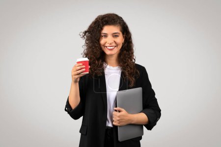 Joven confiada sosteniendo una taza de café y un portátil gris posando sobre un fondo gris, retratando profesionalismo y accesibilidad