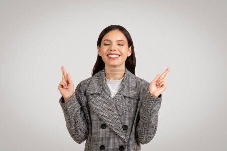 Photo for Hopeful young businesswoman with eyes closed, fingers crossed and beaming smile, symbolizing aspirations and optimism, isolated on clean background, sign of hope and ambition - Royalty Free Image