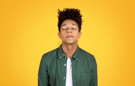 Foto de Un joven negro con una camisa verde se levanta serenamente con los ojos cerrados sobre un fondo amarillo, exudando calma - Imagen libre de derechos