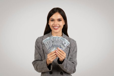 Téléchargez les photos : Femme d'affaires rayonnante présentant propagation de billets de dollar, vêtue d'un blazer à dents de poule classique, symbolisant le succès financier sur fond gris - en image libre de droit