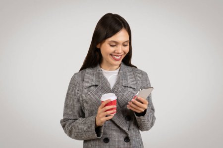 Foto de Ocupada joven empresaria profesional sosteniendo teléfono inteligente y taza de café rojo, interactuando con la tecnología, mientras que en el descanso, vestido con chaqueta houndstooth elegante - Imagen libre de derechos