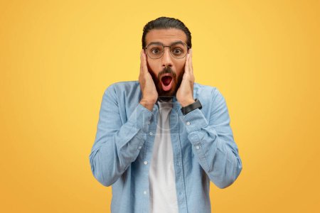 A stunned indian man with glasses holds his cheeks in surprise against a brightly colored yellow backdrop