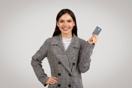 Fröhlicher junger Berufstätiger, der stolz seine Kreditkarte zeigt, gekleidet in einen grauen Hahnenfuß-Blazer, der finanzielles Vertrauen symbolisiert, auf grauem Hintergrund