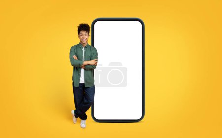 Hombre afroamericano confiado en ropa casual apoyado en una maqueta gigante de teléfonos inteligentes contra un telón de fondo amarillo