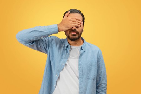 Indian man stands with his hand obscuring his face, implying anonymity or identity protection, set against a vivid yellow background