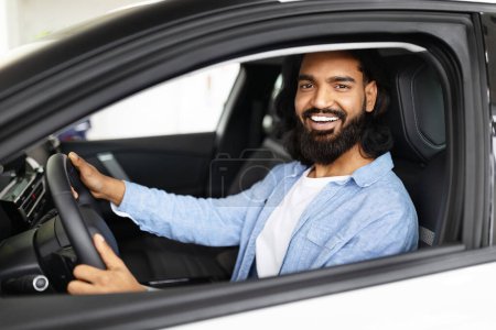 Téléchargez les photos : Un indien barbu joyeux aux cheveux longs est assis au volant d'un véhicule contemporain, regardant la caméra avec un large sourire - en image libre de droit
