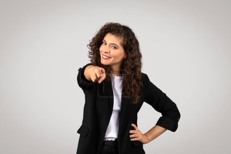Jeune femme confiante avec les cheveux bouclés pointant vers la caméra, portant un blazer sur une chemise décontractée