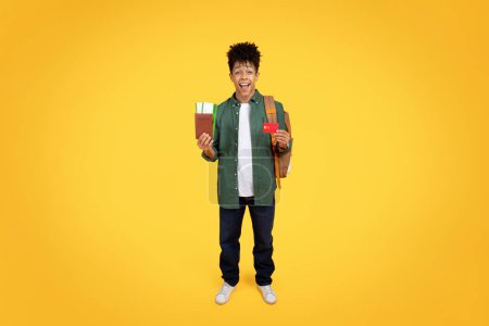 Lächelnder schwarzer Typ mit Rucksack, der einen Reisepass und eine Bordkarte auf gelbem Hintergrund in der Hand hält, Kreditkarte benutzt, Tickets bucht
