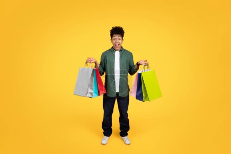 Photo for Excited young black man holding colorful shopping bags and exclaiming with joy against a bright yellow background - Royalty Free Image