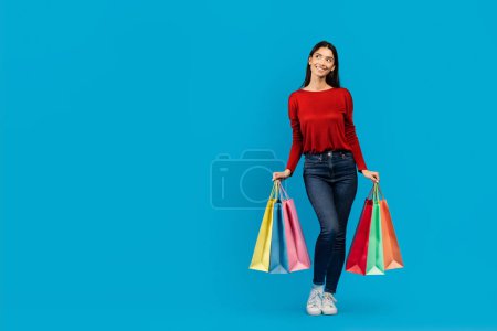 Téléchargez les photos : Une femme portant un haut rouge fait du shopping, tenant plusieurs sacs à provisions dans ses mains et regardant l'espace de copie, heureuse femme debout sur fond bleu - en image libre de droit